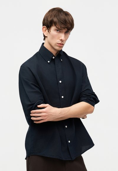 Νεαρός άνδρας με καστανά μαλλιά φορώντας σκοτεινό πουκάμισο με κουμπιά και τα μανίκια σηκωμένα, σταυρώνοντας τα χέρια του μπροστά σε απλό ανοιχτό φόντο.