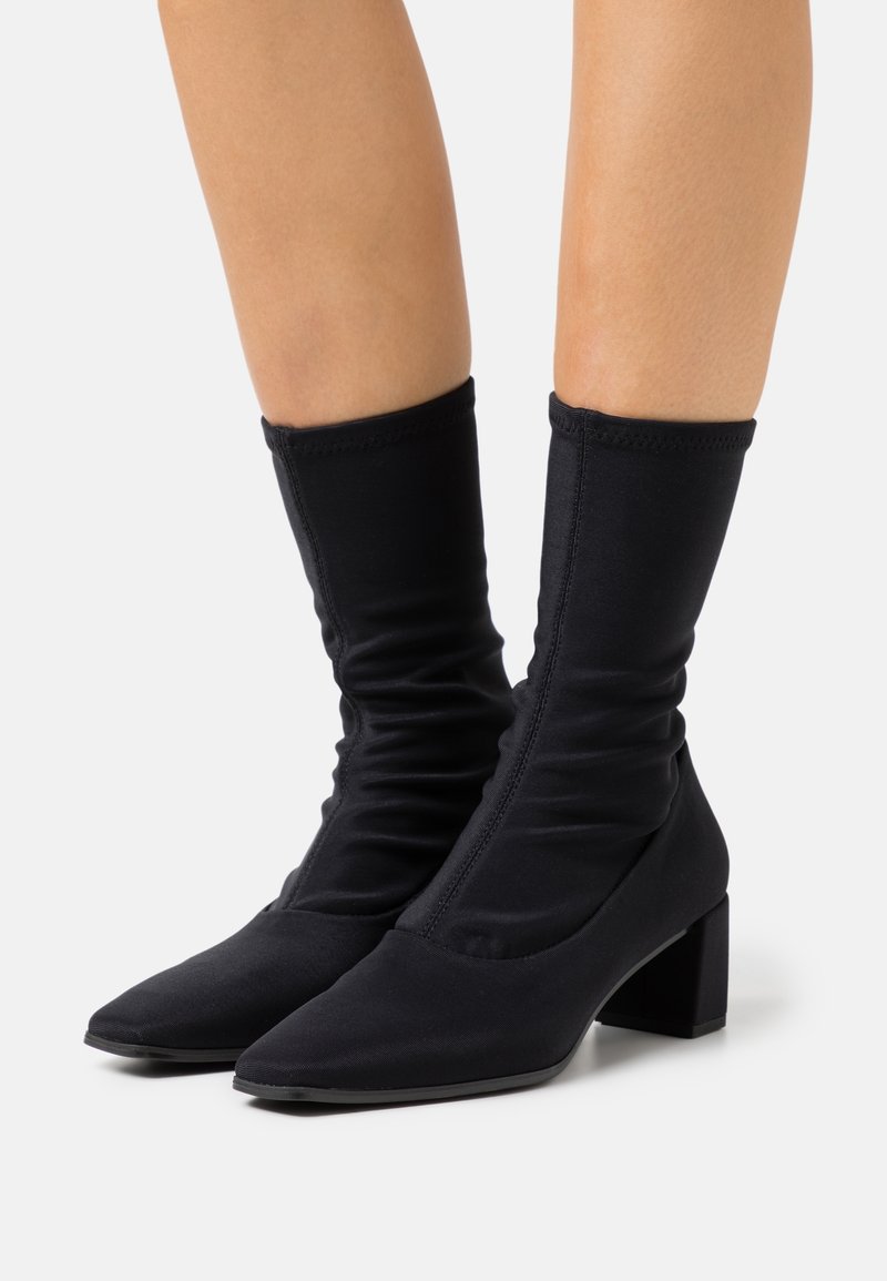 Botas de tobillo de tela negra con un diseño arrugado, punta afilada y tacón cuadrado. La textura suave y el material elástico mejoran el ajuste y la comodidad.