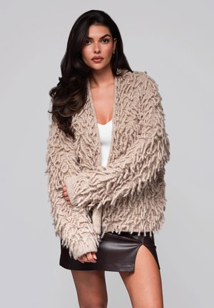 Bežový chunky pletený cardigan s chlupatou texturou, oversized střihem a širokými rukávy. Kombinováno s bílým topem a černou koženou sukní.