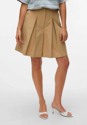 Beige, plisserade shorts med hög midja, tillverkade av en bomullsblandning, har skarpa linjer och en skräddarsydd finish, tillsammans med vita öppna klackskor.