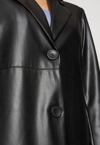 Svart läderrock med stucken krage, två framknappar och en slät textur. Framstycksdetaljer är synliga på kroppen.