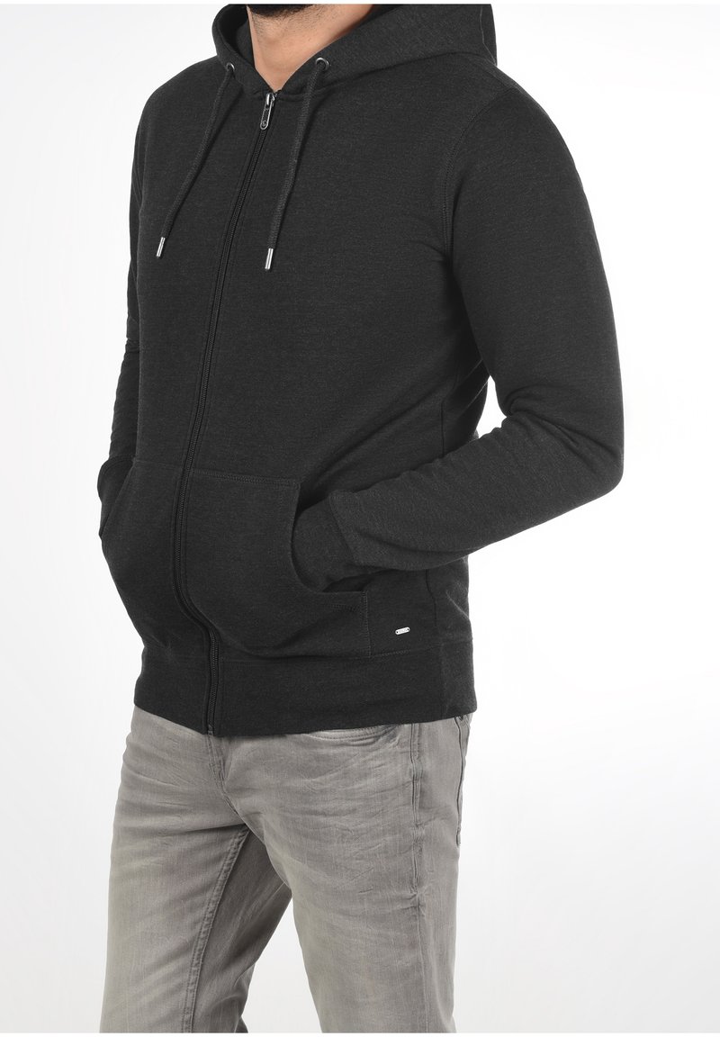 Homme portant un sweat à capuche noir zippé, les mains dans les poches et un jean gris clair usé, debout devant un fond blanc.