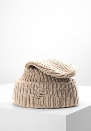Beżowa dzianinowa wełniana czapka z prążkowanym wzorem i podwiniętym mankietem, spoczywająca na białym pedestalzie na jednolitym białym tle.