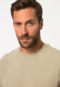 Béžové tričko s kulatým výstřihem a měkkouTexturou, s jemným melanžovým vzorem a žebrovaným límcem. Model má vousy.