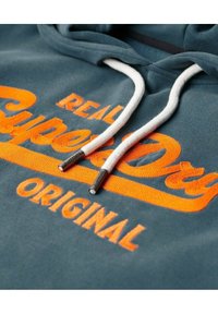Sudadera color azul verdoso con cordones blancos y texto bordado en naranja que dice "REAL Superdry ORIGINAL" en el pecho.