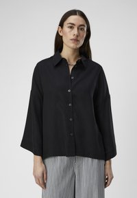 Camisa negra de botones con cuello, fit oversized y mangas anchas. Textura suave, combinada con pantalones de rayas verticales.