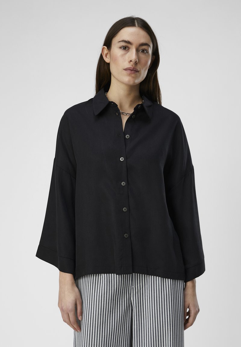 Camisa negra de botones con cuello, fit oversized y mangas anchas. Textura suave, combinada con pantalones de rayas verticales.