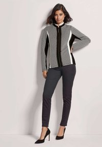 Schwarze und weiße gemusterte Jacke mit hohem Kragen und Frontreißverschluss, kombiniert mit dunklen Hosen und schwarzen High Heels.