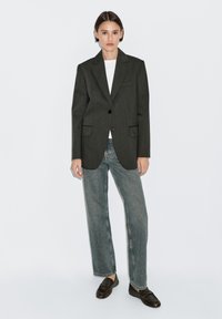 Donkergrijze blazer op maat met uitsnijdingen en voorzakken, gecombineerd met lichtgrijze high-waisted jeans en donkere loafers.