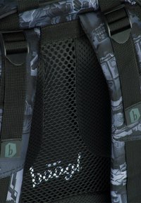Interno di uno zaino con rete nera e cinghie, caratterizzato da un motivo geometrico grigio e un logo di marca bianco. Materiali durevoli e testurizzati.