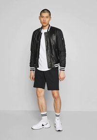 Jeune homme debout, portant une veste bomber noire et blanche, un t-shirt blanc, un short noir, des chaussettes rayées et des baskets blanches Nike.