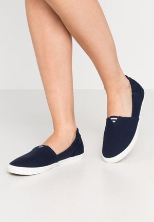 Ben iført mørkeblå slip-on lærredsko med hvide gummisåler på en lysegrå gulv.
