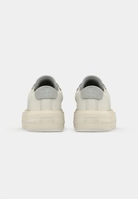 Un paio di sneakers Converse All Star off-white viste da dietro, con suole spesse e linguette grigie sui talloni, su uno sfondo neutro.