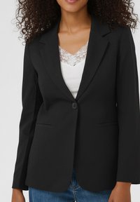 Blazer noir à un seul bouton, col en pointe, et deux poches avant. Tissu texturé, silhouette ajustée, porté sur un haut en dentelle blanche.