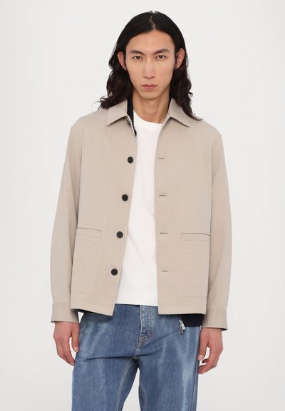 Jachetă bej deschis din bumbac, cu guler, nasturi în față și două buzunare laterale. Purtați-o peste o cămașă albă cu pantaloni din denim albaștri.