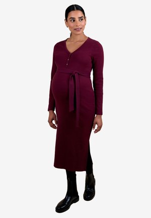 Gravid kvinde iført en langærmet, burgunderfarvet ribstrikket kjole med taljebånd, sorte leggings og sorte ankelstøvler, stående og kigger til siden.