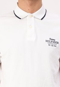 Hvit polo skjorte med teksturert stoff, marineblå krage og en liten marineblå brodert logo på venstre bryst. Har to knapper.