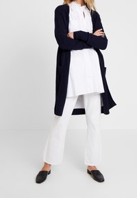 Mujer vestida con un cárdigan azul marino sobre una camisa blanca de botones, pantalones anchos blancos y zapatos negros sin cordones, de pie con los brazos cruzados.