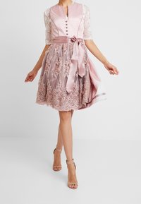 Robe rose avec des manches en dentelle, présentant un corsage ajusté, des boutons, et une jupe évasée ornée de broderies florales et d'une ceinture en satin.