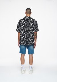 Camisetta nera a maniche corte con motivo floreale bianco, abbinata a pantaloni shorts in denim blu. Il modello indossa sneakers bianche e calzini a fantasia.