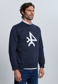Sudadera azul marino de algodón, con un gran logo gráfico blanco en el frente, cuello redondo y puños acanalados. Se lleva con jeans de mezclilla.