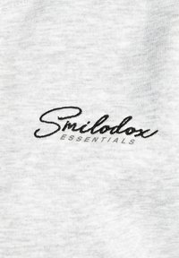 Tessuto grigio con una texture liscia, con 'Smilodox' ricamato in corsivo nero sopra 'ESSENTIALS' in un font più semplice.