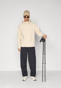 Beige fleece pullover with high collar, dark gray cargo pants, cream sneakers, and hiking poles. Accessories include a beige beanie and sunglasses.
