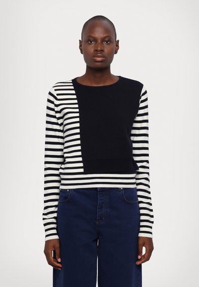 Pull rayé avec des rayures horizontales noires et blanches sur les manches, corps noir uni, col rond et coupe décontractée. Associé à un pantalon bleu foncé.