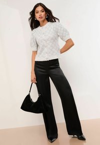 Krátkozrčný, svetlošedý pletený kardigan s prešívaným vzorom a veľkými gombíkmi, skombinovaný s čiernymi saténovými nohavicami so zvýšeným pásom a čiernou kabelkou.