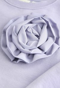 Lilac džemperis ar slāņotu auduma rozes aplikāciju priekšpusē, ar mīkstu tekstūru un gludu apdari.