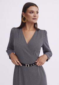 Czarna i biała w paski sukienka w stylu wrap z długimi rękawami, głębokim dekoltem w kształcie V i czarnym paskiem z złotą klamrą. Wyraźne złote kolczyki w kształcie kół.