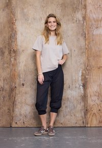Jonge vrouw die lacht, staande tegen een houten muur, draagt een beige t-shirt, zwarte cropped broek en bruine sneakers met witte strepen.
