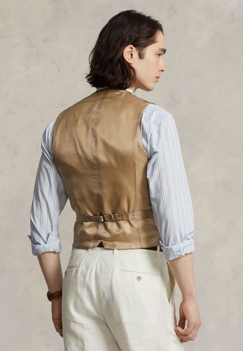 Gilet en satin beige avec une texture lisse, doté d'une boucle à l'arrière pour l'ajustement. Porté par-dessus une chemise à rayures bleu clair, manches retroussées.