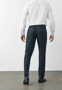 Pantaloni negri comoști, cu un finisaj texturat, având două buzunare la spate, asortați cu o cămașă albă. Pantofi din piele negri vizibili.
