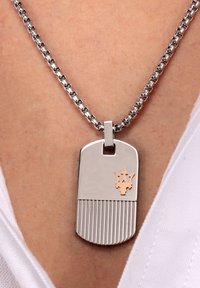 Collana con pendente in argento a forma di dog tag, con fondo rigato e logo a tridente dorato; presenta una catena metallica con anelli intrecciati.