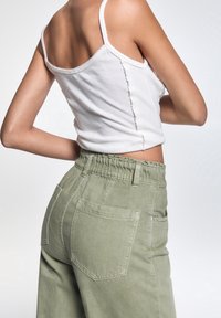 Mujer vestida con una blusa sin mangas blanca y pantalones verdes olivo de cintura alta, vista desde atrás a la altura de la cintura sobre un fondo sencillo.