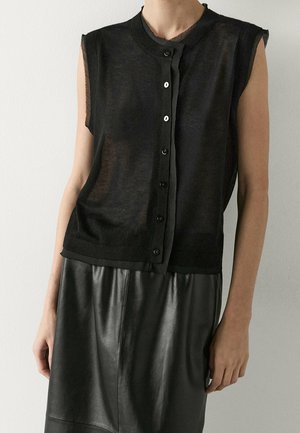 Personne portant un haut noir transparent sans manches à boutons et une jupe en cuir noire, debout devant un mur clair uni.