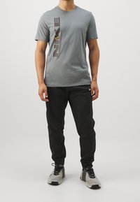 Grå bomullst-shirt med korta ärmar, med en stor vertikal svart Nike-logotyp och en gul swoosh. Kombinerad med svarta byxor och grå sneakers.