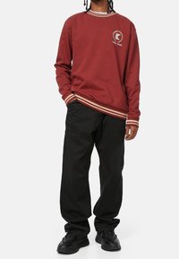Persona che indossa una felpa bordeaux con polsini a righe e pantaloni neri, in piedi con una mano in tasca su uno sfondo bianco.