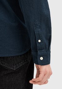 Námořnická strukturovaná košile s knoflíkovými manžetami se dvěma bílými knoflíky, v kombinaci s tmavými džíny, zdůrazňující detail řemeslného zpracování.