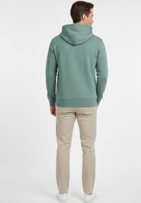 Sudadera con capucha, verde claro, tela texturizada, puños y dobladillo acanalados. Combinada con pantalones beige claro y zapatos blancos, vista desde atrás.