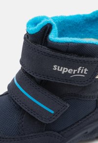Botte pour enfants bleu foncé avec une sangle velcro, en tissu texturé et doublure en polaire bleu clair. Possède une rayure d'accent bleu et un logo.
