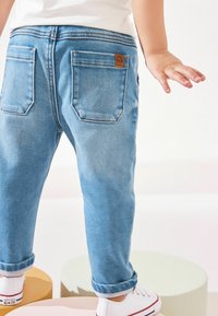 Jean en denim bleu clair avec ourlets roulés, deux poches arrière et une pièce en cuir beige sur la taille. Des baskets blanches complètent la tenue.