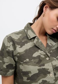 Camicia a maniche corte verde oliva con motivo mimetico, dotata di colletto, chiusura con bottoni e tessuto dalla texture morbida.