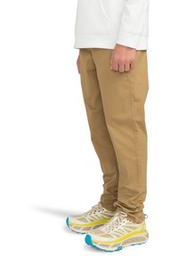 Khakifärgade bomullsbukser kombinerat med beige och gula sportskor, med en texturerad sula och blå accenter. Vit sweatshirt synlig på toppen.