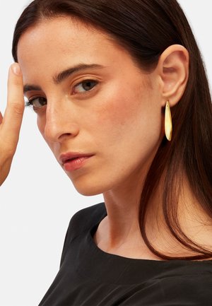 Gouden, langwerpige druppeloorbellen hangen aan het oor van een model en weerkaatsen het licht met een gladde, gepolijste oppervlakte.