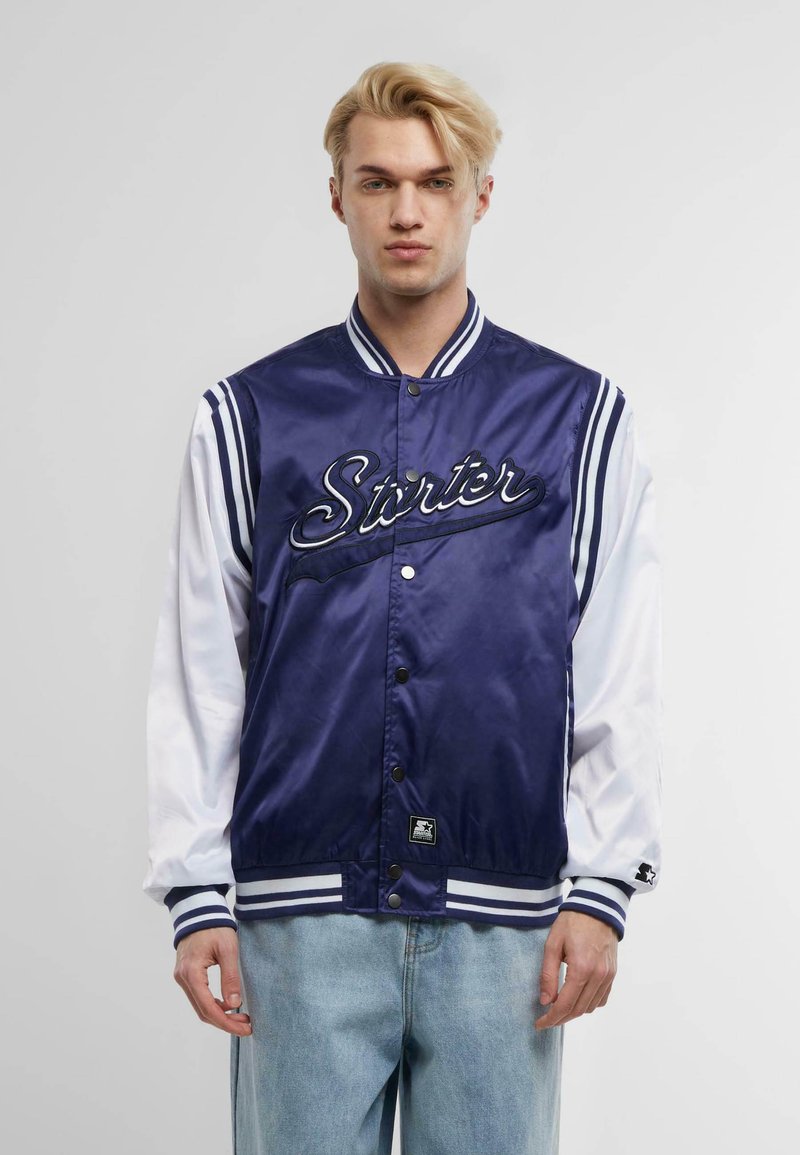 Navy satin varsity jacket with white sleeves, featuring "Starter" embroidered on the front, striped cuffs, and snap buttons. Denim pants complete the look.