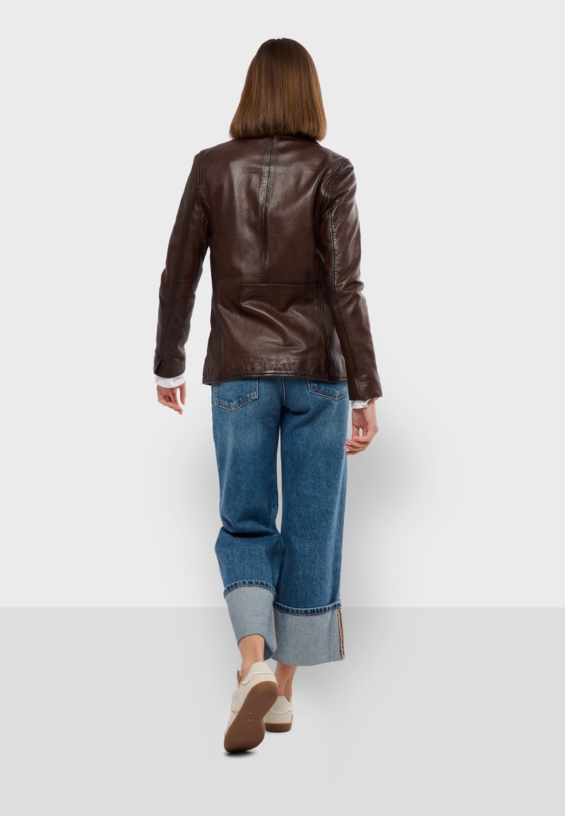 Giacca di pelle marrone con un design aderente, texture liscia e cuciture distintive. Abbinata a jeans a gamba ampia azzurri e scarpe bianche.