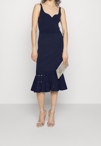 Robe ajustée bleu marine avec un décolleté cœur, ourlet évasé comportant des découpes, assortie à des talons argentés à lanières et une pochette.