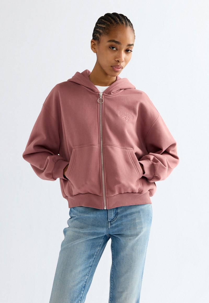 Jeune femme aux cheveux tressés portant un sweat à capuche zippé rose poudré et un jean bleu clair, les mains dans les poches du sweat à capuche, expression neutre.